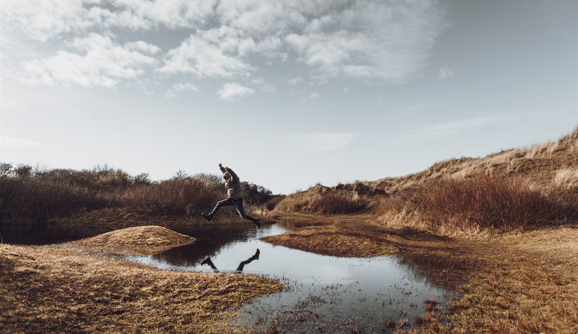 Onder een grijze, bewolkte lucht, springt een man over een grote plas heen met zijn armen in de lucht